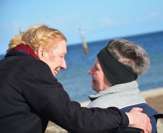 Eine Frau lächelt eine Frau im Rollstuhl an. Beide sind am Meer.