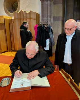 Ein Mann unterschreibt in einem großen Buch, während zwei weitere Männer in einer Kirche zusehen.