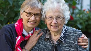 Zwei ältere Frauen sitzen eng beieinander, eine trägt ein schwarz-weiß gemustertes Oberteil, die andere einen roten Schal mit weißem Muster.