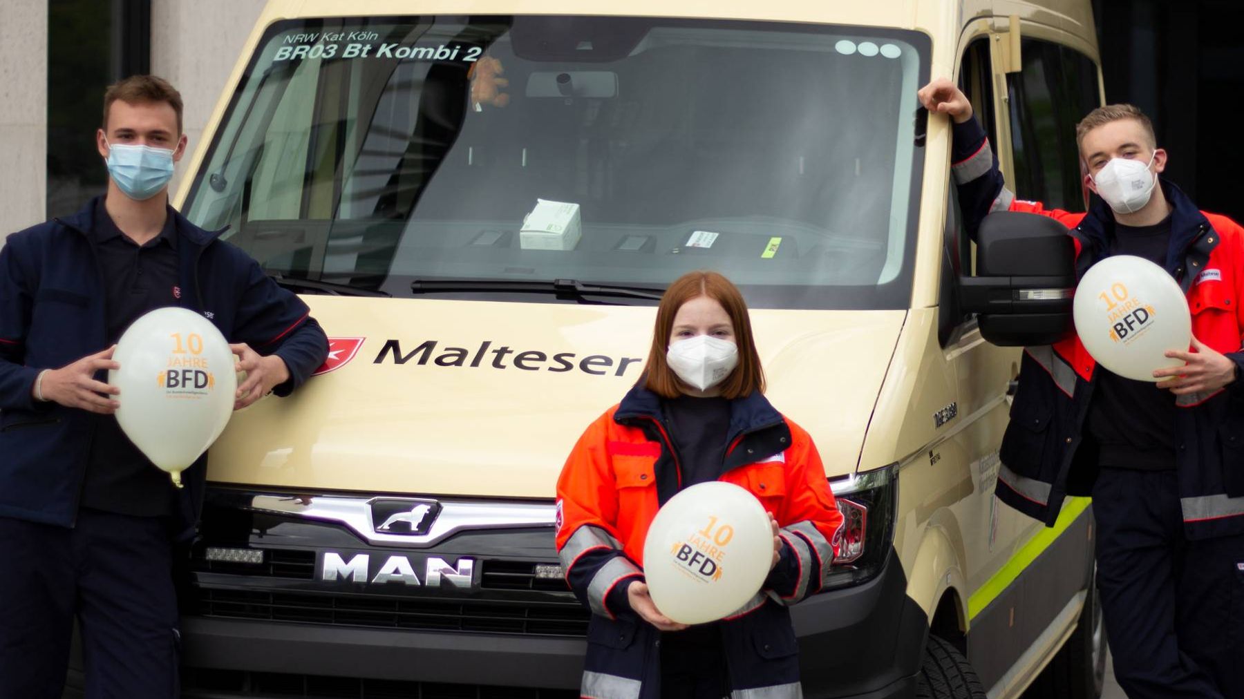 Eine junge Frau und zwei junge Männer halten Luftballons vor einem Kleinbus in die Höhe