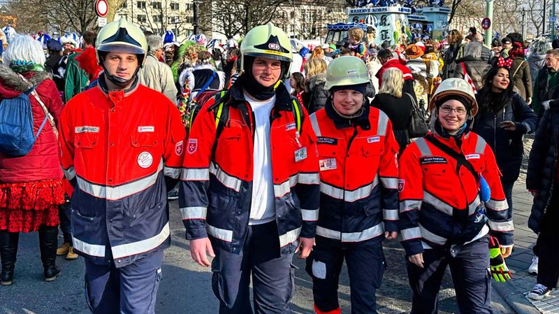 Drei Männer und eine Frau in Einsatzkleidung und Helm laufen auf einer asphaltierten Straße entlang. 