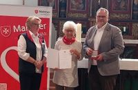 Eine Frau zeigt eine Urkunde, neben ihr stehen ein Mann und eine Frau in Malteser Kleidung.