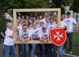 Eine Gruppe von Menschen mit Malteser Social Day T-Shirts halten einen großen Holzrahmen