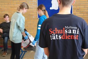Junge von hinten mit großer Tshirt Aufschrift mit den Worten Malteser Schulsanitätsdienst. Vor ihm stehen zwei Schulsanitäterinnen, die einem fiktiven Patienten, der auf einem Stuhl sitzt, den Blutdruck messen.