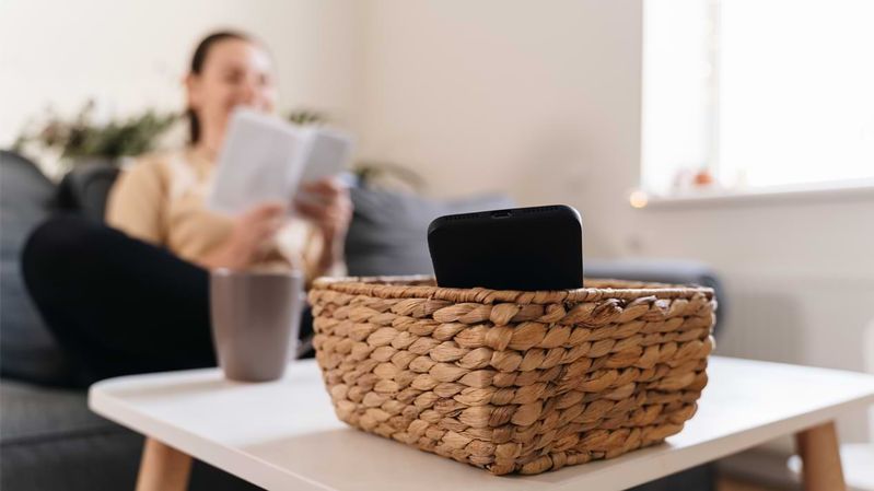 Eine Frau im Hintergrund liest ein Buch. Im Vordergrund ein Smartphone in einem Korb. 