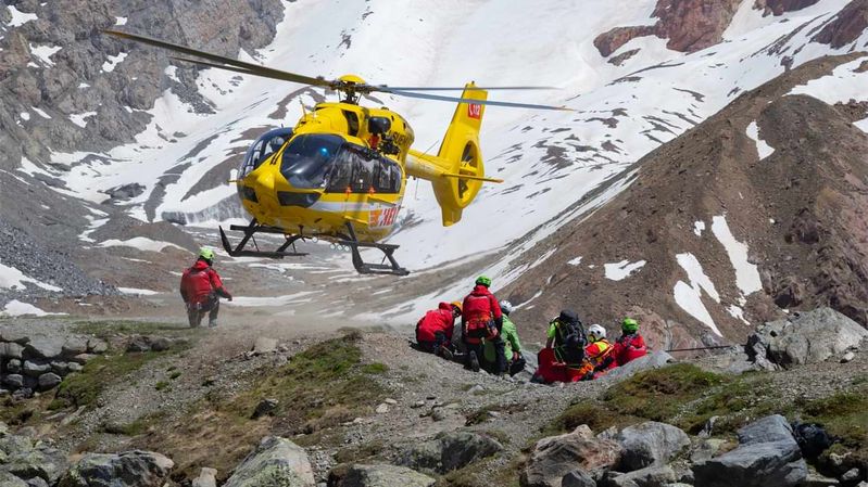 Angehörige der Bergwacht in roter Einsatzkleidung und mit grünen Helmen unter einem gelben Hubschrauber in einer alpinen Landschaft.