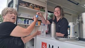 Eine Frau reicht einer anderen eine kleine Flasche mit einem Pflegemittel.