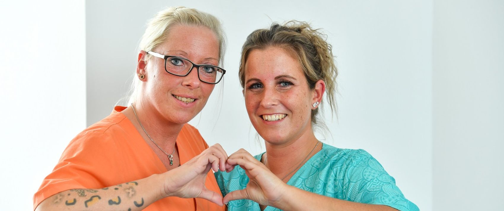 Zwei Frauen formen gemeinsam mit den Händen ein Herz und lächeln dabei in die Kamera