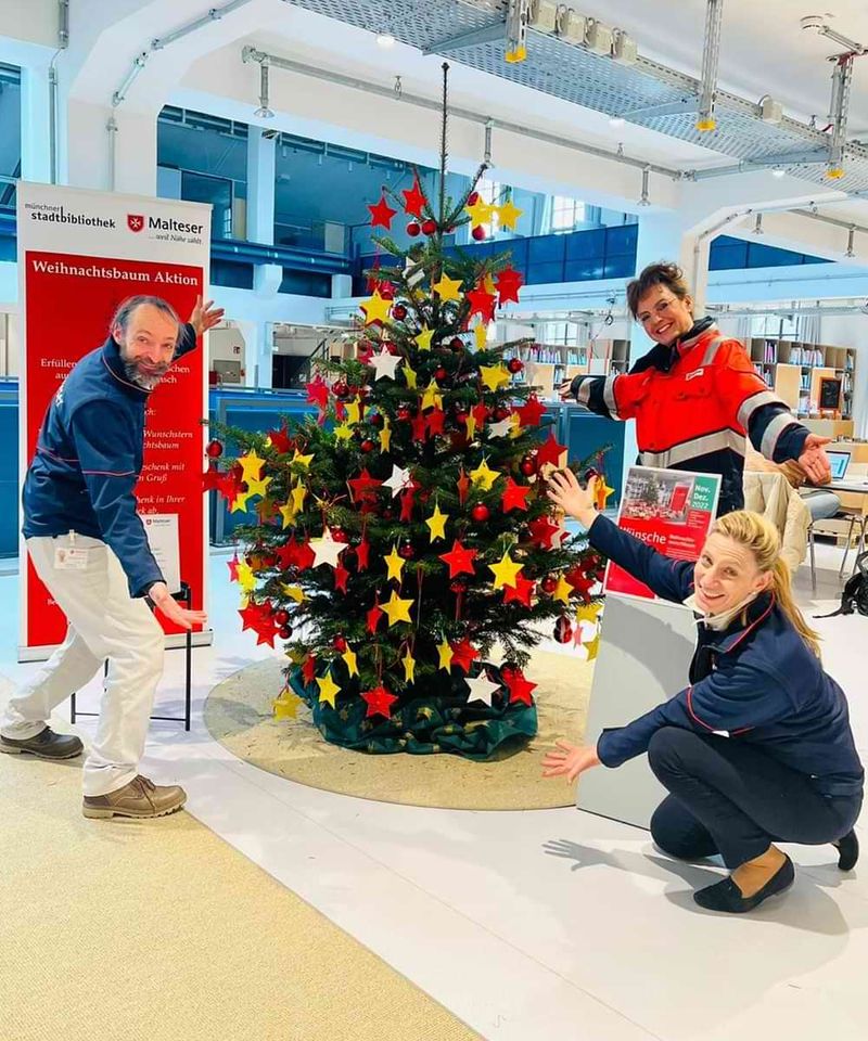 Ein Weihnachtsbaum, behängt mit vielen Sternen, steht in einer Bibliothek und wird von drei Mitgliedern der Malteser präsentiert.