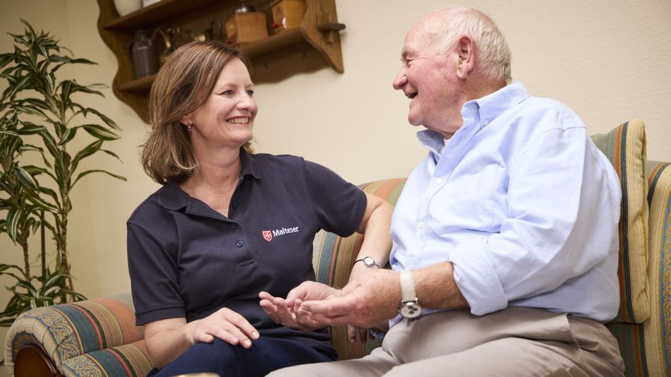 Eine lächelnde Frau und ein älterer Mann sitzen auf einem Sofa und unterhalten sich.