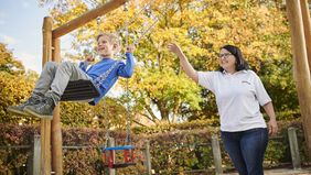 Ein Kind sitzt auf einer Schaukel. Ein Frau begleitet dieses dabei.
