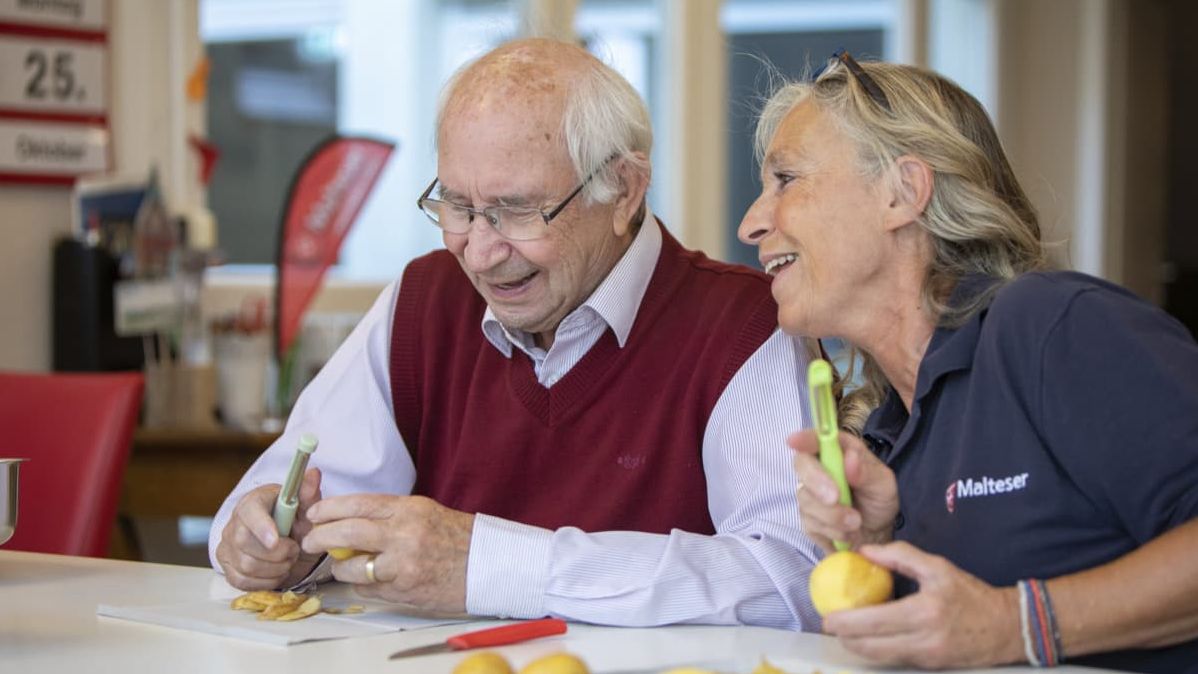 Älterer Mann und Helferin der Malteser schälen fröhlich Kartoffeln