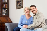 Eine ältere Dame mit Hausnotruf-Knopf sitzt mit ihrem Sohn auf dem Sofa und lacht