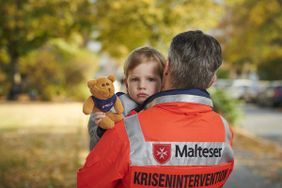 Ein Mann in Malteser Jacke trägt ein Kind mit Teddy auf dem Arm