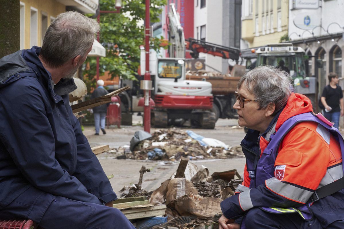 Eine Frau kniet vor einem Mann. Im Hintergrund Aufräumarbeiten.