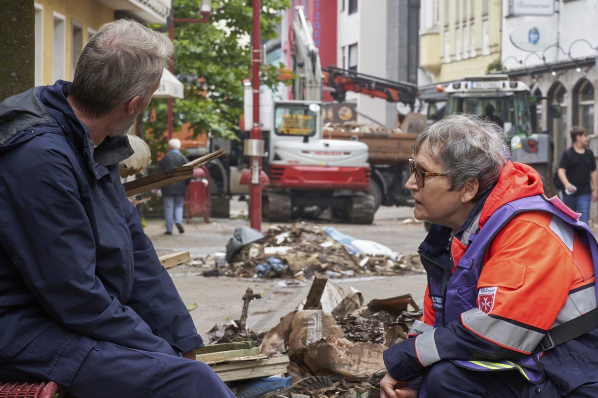Eine Frau kniet vor einem Mann. Im Hintergrund Aufräumarbeiten.