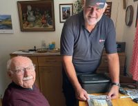 Der Senior sitzt am Tisch, während der Malteser Menüservice-Mitarbeiter die Menüschale für ihn öffnet.