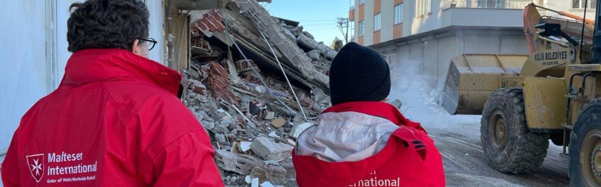 Zwei Menschen mit roten Jacken vor einem zerstörten Haus.