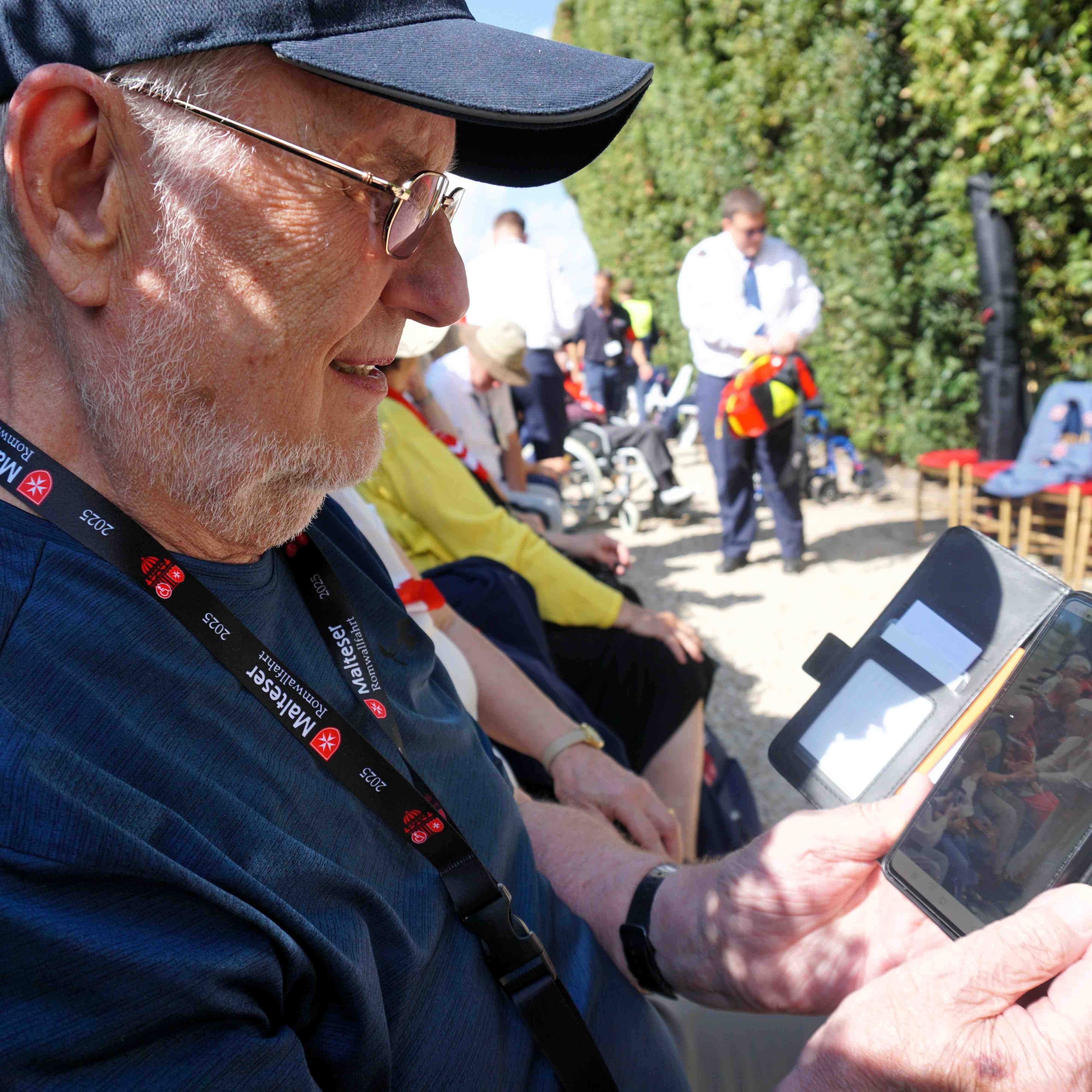 Mann mit Baseballkappe hält Tablet, im Hintergrund weitere Personen auf einem Weg neben grünen Büschen.