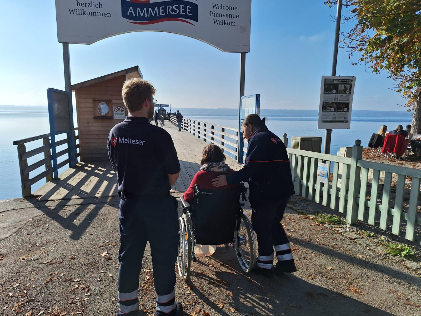 2 Malteser Ehrenamtliche und eine Person im Rollstuhl vor einem Ammersee Dampfersteg