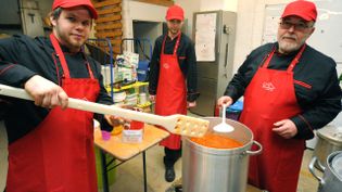 3 Köche stehen in einer Feldküche vor einem sehr großen Topf.