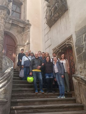 Eine Gruppe von Teilnehmenden steht auf den Stufen eines reich verzierten historischen Gebäudes und posiert für ein gemeinsames Erinnerungsfoto. Die kunstvoll gestaltete Fassade und die Treppenanlage verleihen der Szene einen besonderen Rahmen.