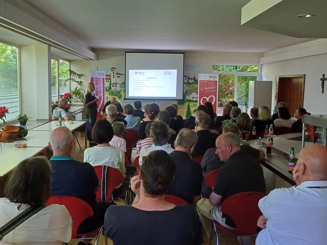 Im Vordergrund sitzen zahlreiche Mitarbeitende in Stuhlreihen, während auf einer Leinwand die Tagesordnungspunkte dargestellt werden.