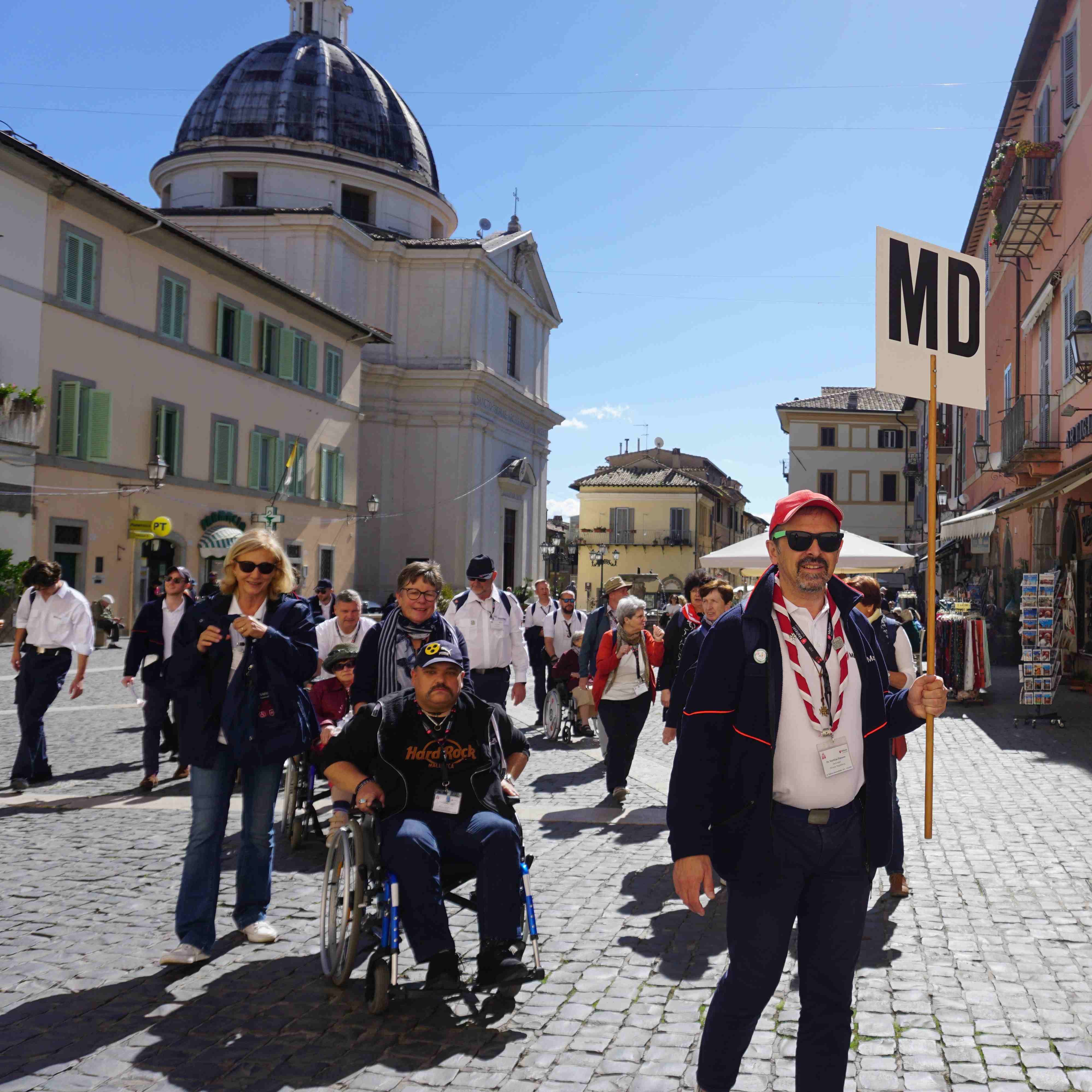 Menschenmenge auf gepflastertem Platz in Castel Gandolfo, im Hintergrund Kuppel eines historischen Gebäudes und mehrstöckige Häuser.