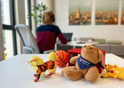 Teddy auf dem Tisch. Im Hintergrund arbeitet eine Frau am Laptop Frau am SchreibtischFoto: Malteser, C. Düpjohann