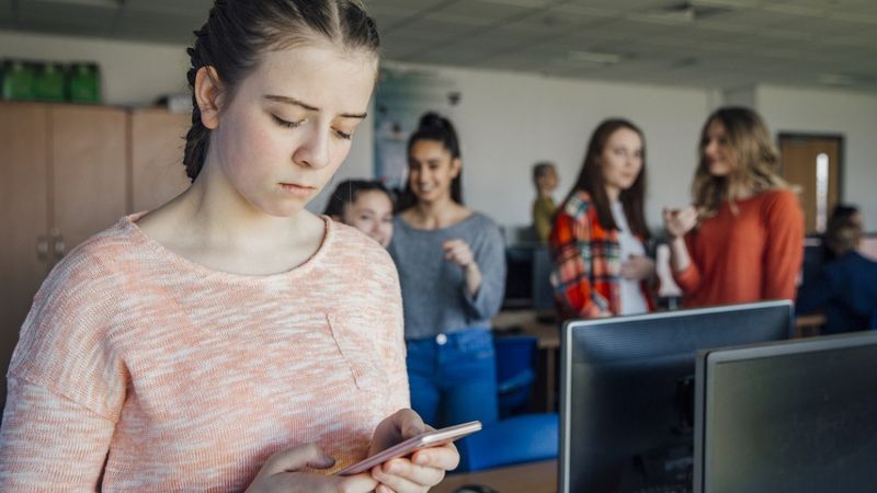 Ein Mädchen in einem Klassenzimmer schaut auf ein Smartphone; im Hintergrund stehen weitere Schülerinnen