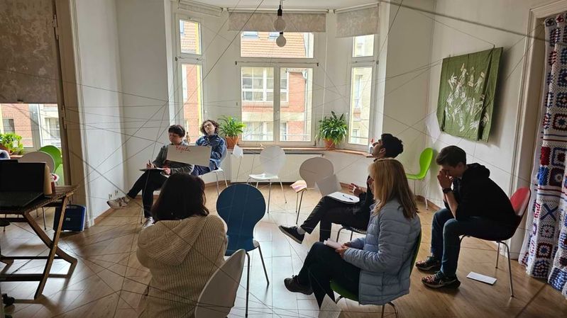 Junge Menschen sitzen auf Stühlen in einem Raum, in dem Fäden kreuz und quer gespannt sind.