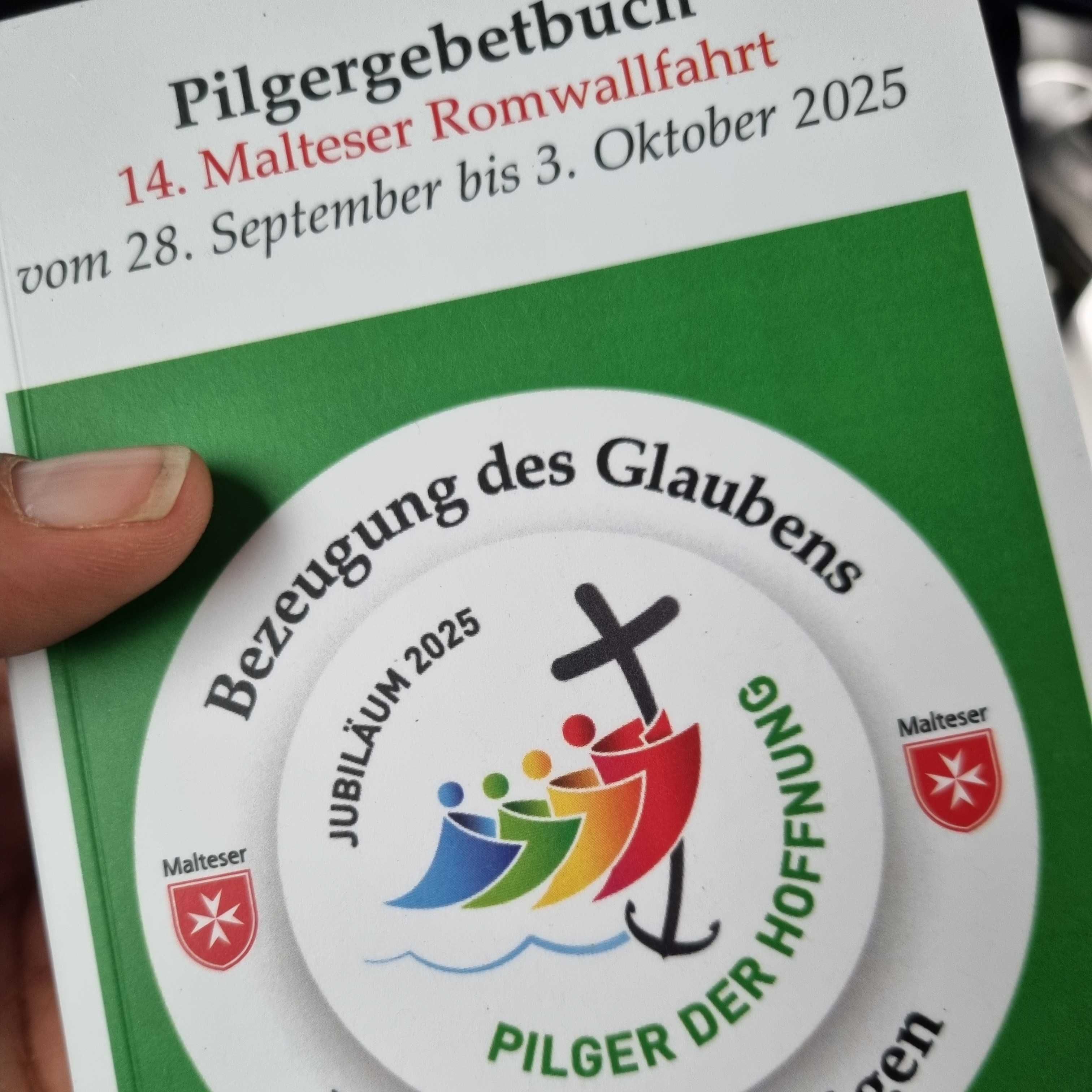 Hand hält Pilgergebetbuch mit Titel '14. Malteser Romwallfahrt vom 28. September bis 3. Oktober 2025'. Auf Buchdeckel runder Aufkleber mit bunten stilisierten Figuren, Kreuz, Anker und Text 'Bezeugung des Glaubens', 'Jubiläum 2025', 'Pilger der Hoffnung'.