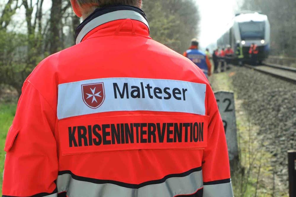 Die Rückenansicht eines Malteser Helfers. Im Hintergrund sieht man ein Zugünglück.
