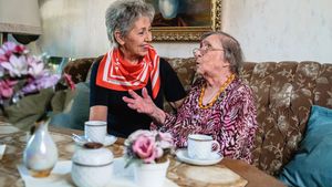 Zwei ältere Frauen sitzen in einem Wohnzimmer auf einem Sofa vor Kaffeetassen.