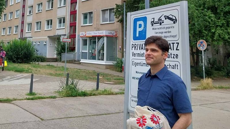 Ein Mann in einem blauen Hemd mit einem Jutebeutel vor einem Schild, das auf einen Privatparkplatz hinweist.
