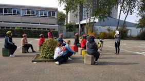 Die Frauen sitzen draußen auf dem Schulhof auf Steinen und vorne erklärt die Trainerin die Bedeutung eines Verkehrsschildes, welches sie hoch hält. 