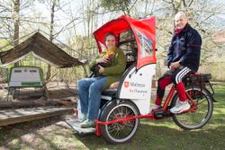 Auf der Fahrradrikscha sitzt vorne eine Gästin mit einem Huhn. Auf dem Sattel sitzt eine ehrenamtliche Pilotin