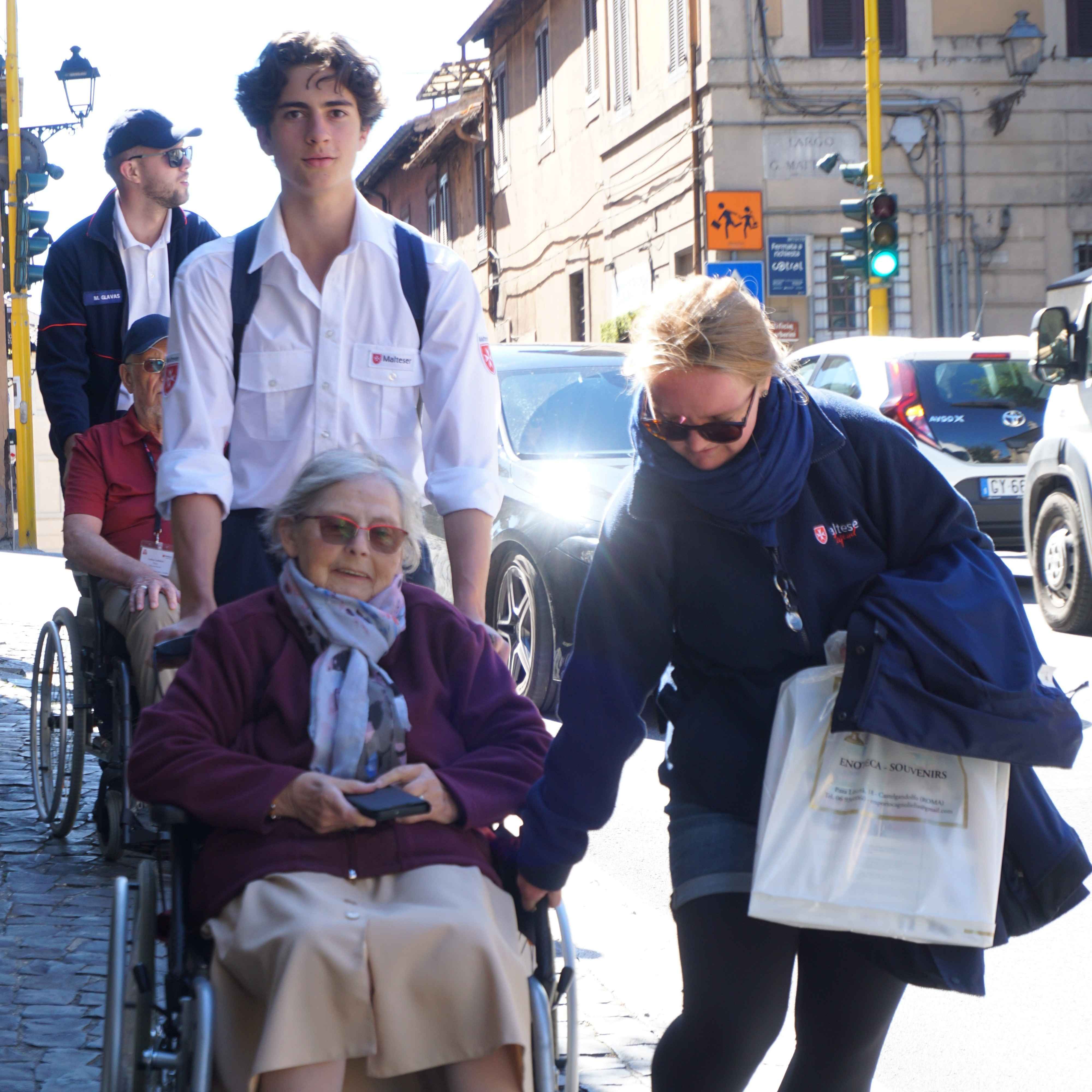 Frau schiebt ältere Person im Rollstuhl auf einer Straße, beide tragen Jacken, im Hintergrund weitere Personen und Autos sichtbar.