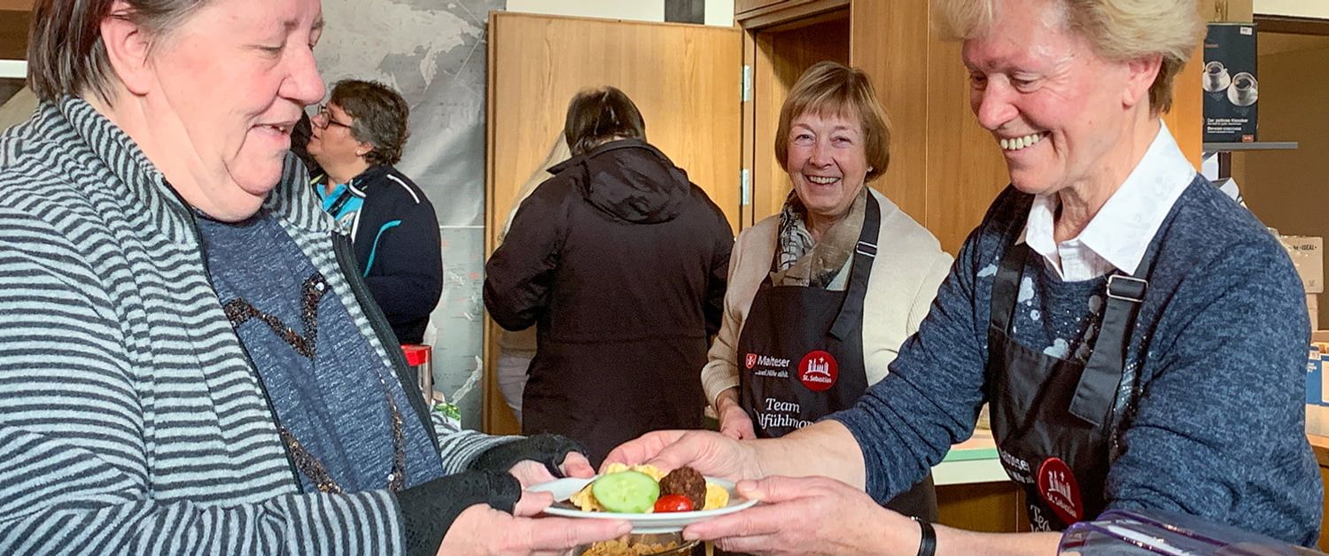 Beim Malteser Wohlfühlmorgen reicht eine Helferin einer Frau Essen über den Tisch.