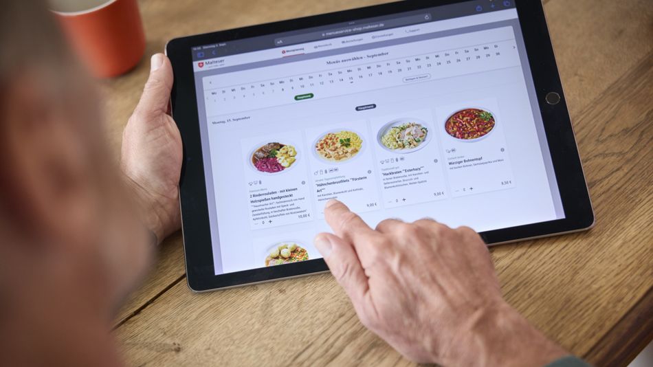 Die Hand eines älteren Mannes wählt auf einem Tablet eines von fünf abgebildeten Gerichten auf einer Website aus.
