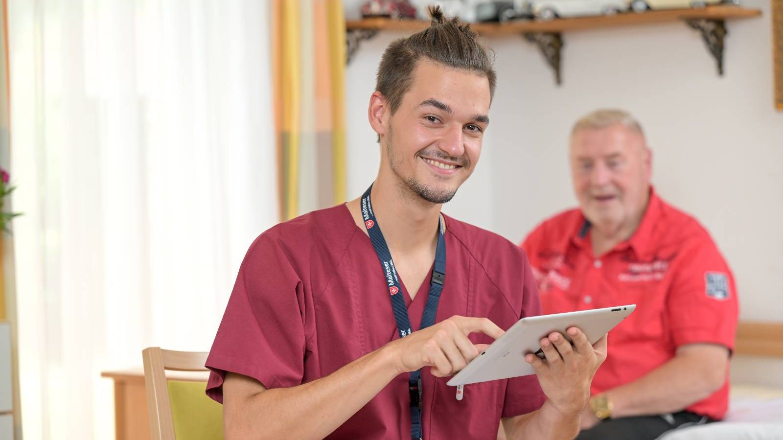 Pfleger der Malteser mit einem Tablet, im Hintergrund ein Patient