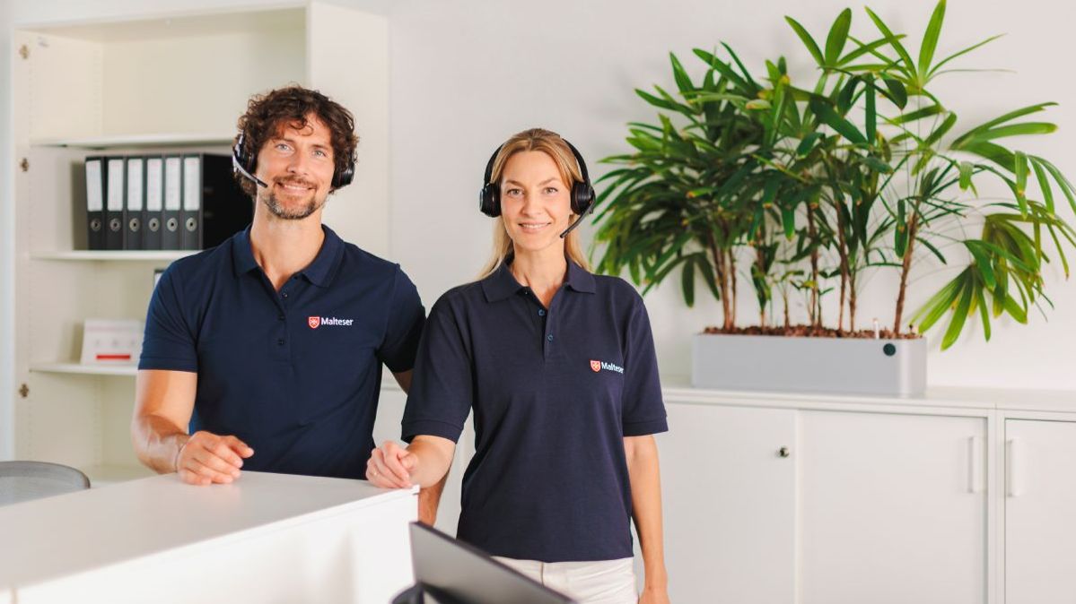 Ein Mitarbeiter und eine Mitarbeiterin der Malteser Hausnotruf Zentrale stehen in einem Büro und tragen ein Headset.