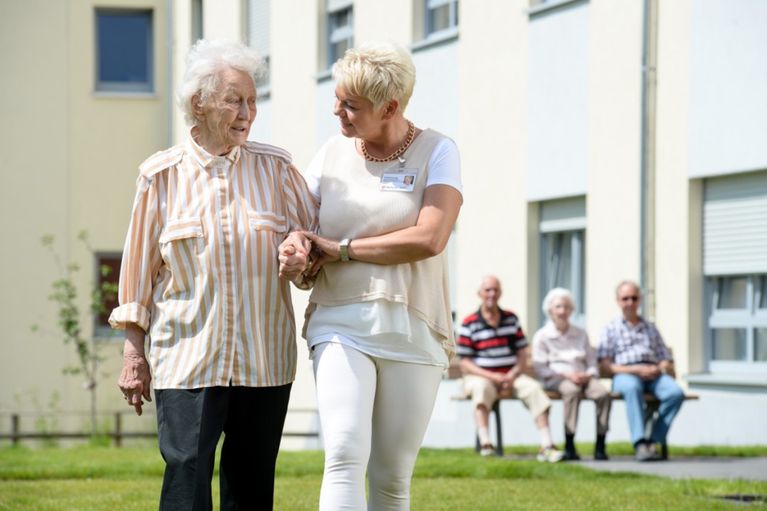 Eine ältere Person wird auf dem Außengelände einer Pflegeeinrichtung von einer Betreuungskraft beim Gehen begleitet, während weitere Bewohnerinnen und Bewohner im Hintergrund sitzen.