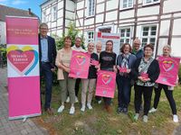 Eine Gruppe von zehn Menschen steht im Freien vor einem Fachwerkhaus mit weiß-brauner Fassade. Sie posieren für ein Gruppenfoto. Links im Bild steht ein großes, aufstellbares Banner mit bunten Herzformen und dem Text „Mensch Hamm! Lebe Freiheit! Mein Herz schlägt für Demokratie“. Einige Personen halten pinkfarbene Plakate oder Flyer mit ähnlichem Design in der Hand. Auf den Plakaten steht: „Wir sehen Dich und hören Dir zu!“ und „Wir sammeln DEINE Herzenswünsche für die Politik der Stadt Hamm“. Die Gruppe besteht aus Männern und Frauen verschiedenen Alters. Alle Personen wirken freundlich und lächeln in die Kamera. Die Stimmung ist positiv und gemeinschaftlich. Im Hintergrund ist ein weiteres Schild mit der Aufschrift „Geschäftsstelle“ zu sehen.