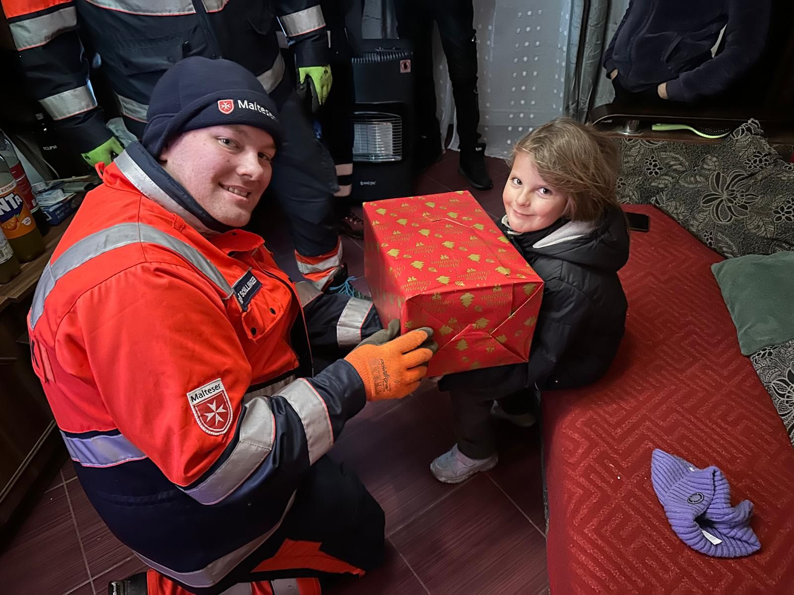 Mann in Malteser Kleidung überreicht einem Mädchen ein rot eingepacktes Weihnachtspaket