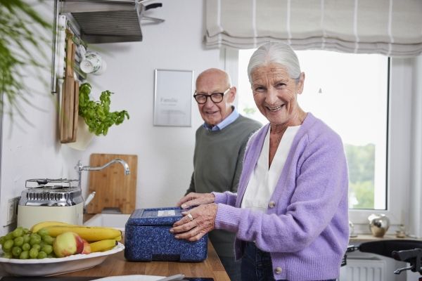 Eine ältere Frau packt ein Menüservice-Essen aus der Thermobox, im Hintergrund steht ein älterer Mann
