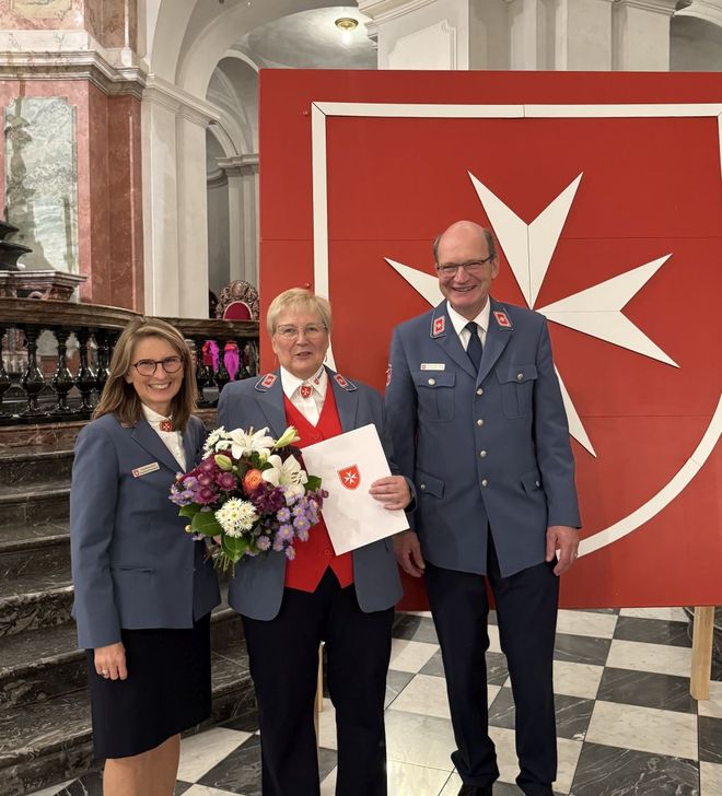 Drei Personen in blauen Uniformen stehen in einer Kirche, die mittlere hält einen Blumenstrauß und ein Dokument.