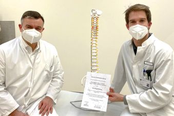 Zwei Ärzte im weißen Kittel sitzen neben einem Wirbelsäulenmodell und halten ein Zertifikat.