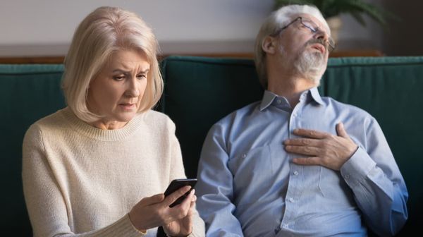 Neben einer Frau mit Handy in der Hand sitzt ohnmächtig ein älterer Mann und fasst sich ans Herz