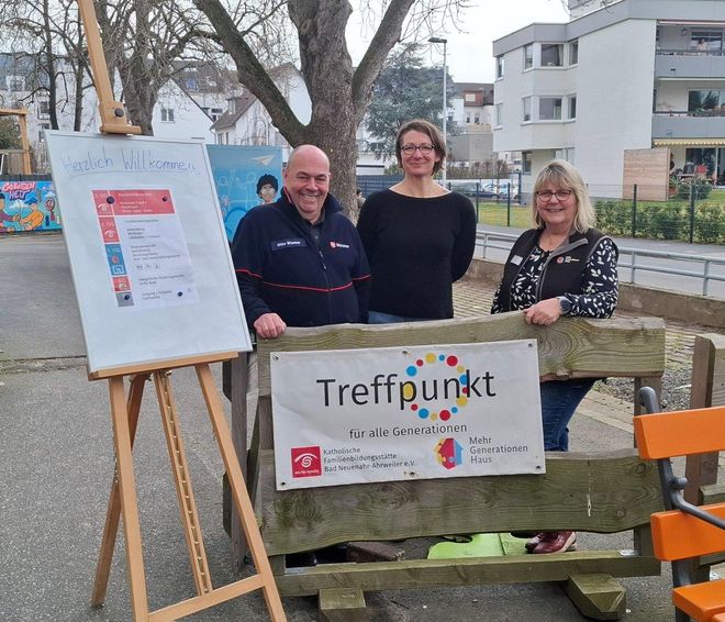 Ein Malteser-Ehrenamtler steht mit zwei Kooperationspartnerinnen im Freien an einem Schild mit dem Titel "Treffpunkt"
